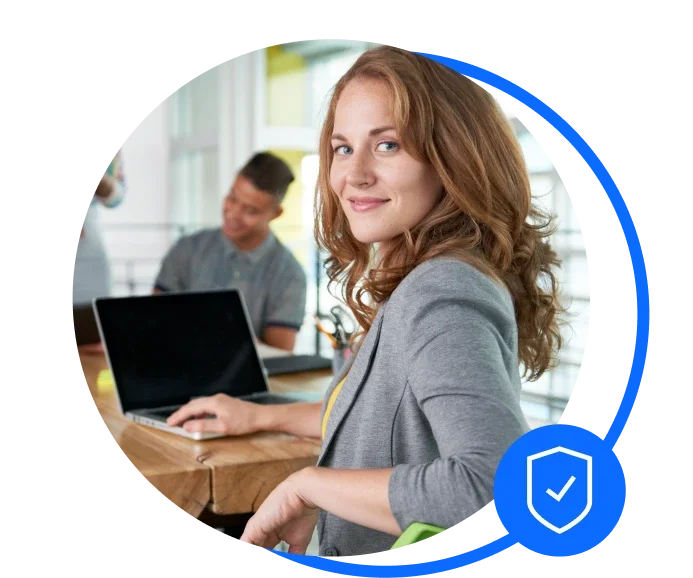 Brunette woman wearing a grey blazer is sat down at a wooden desk with her Mac open. She's turning towards the camera smiling. In the background you can see another male colleague working.